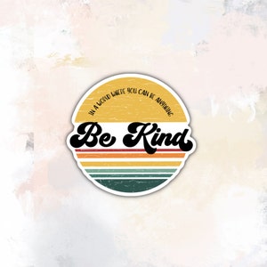 May include: A yellow and orange retro-style sticker with a distressed look. The sticker has a white border and says "In a world where you can be anything, Be Kind".