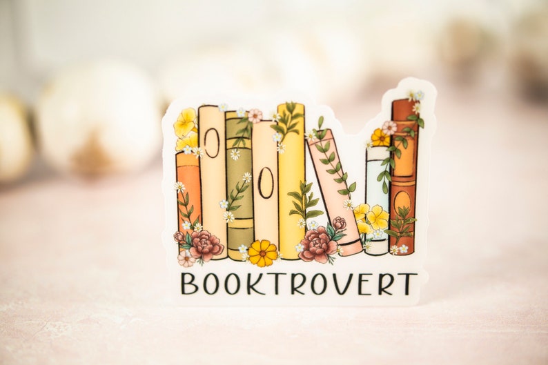 Pu&ograve; includere: Un adesivo con una pila di libri con accenti floreali e il testo "BOOKTROVERT" stampato sotto.