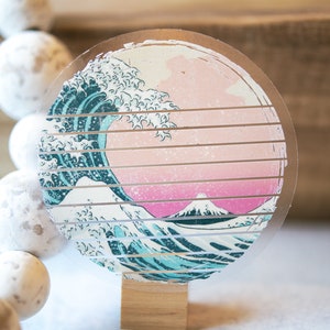 May include: A round, clear sticker with a graphic of a blue and white wave crashing over a pink and white mountain. The sticker is on a wooden background.