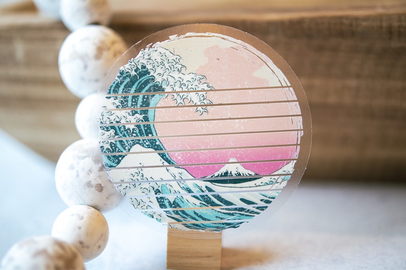 May include: A round, clear sticker with a graphic of a blue and white wave crashing over a pink and white mountain. The sticker is on a wooden background.