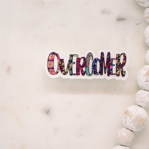 Puede incluir: Una pegatina colorida con la palabra "Overcomer" escrita en una fuente decorativa. La pegatina est&aacute; sobre una superficie de m&aacute;rmol blanco.