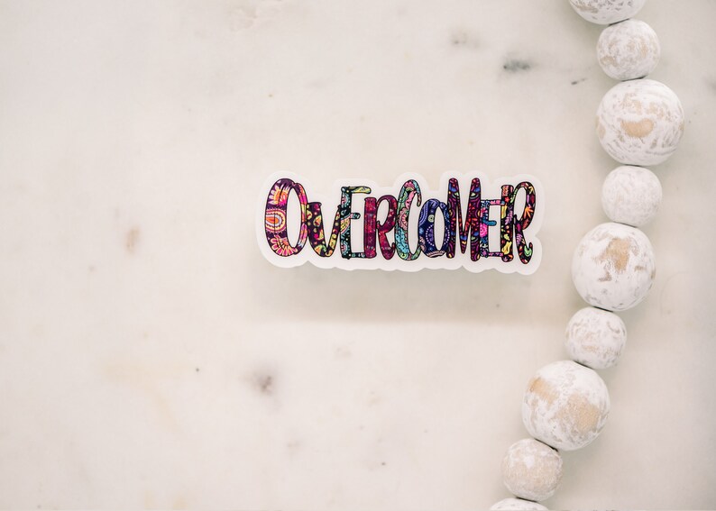 Puede incluir: Una pegatina colorida con la palabra "Overcomer" escrita en una fuente decorativa. La pegatina est&aacute; sobre una superficie de m&aacute;rmol blanco.