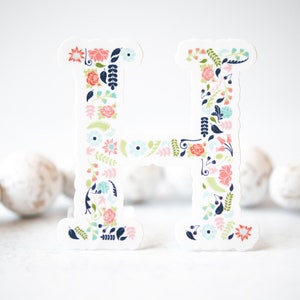 May include: A white paper cutout of the letter H decorated with a floral pattern in shades of pink, blue, green, and navy.
