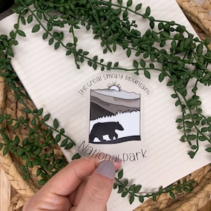 Puede incluir: Una pegatina transparente con una silueta de oso negro caminando frente a una cordillera con un sol saliendo detr&aacute;s de las monta&ntilde;as. El texto "The Great Smoky Mountains National Park" est&aacute; impreso alrededor de la imagen.