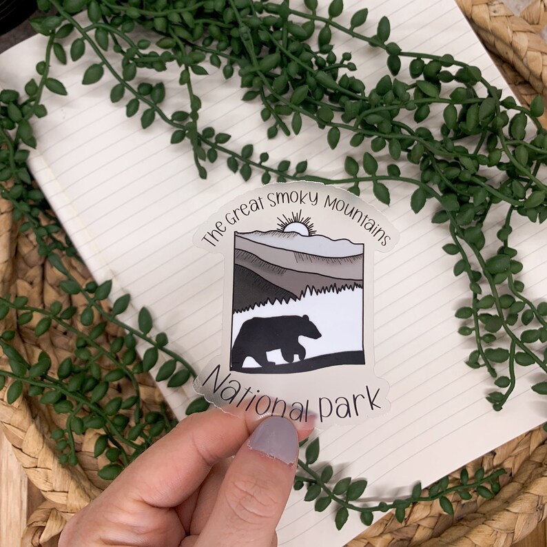Puede incluir: Una pegatina transparente con una silueta de oso negro caminando frente a una cordillera con un sol saliendo detr&aacute;s de las monta&ntilde;as. El texto "The Great Smoky Mountains National Park" est&aacute; impreso alrededor de la imagen.