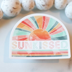 May include: A sticker featuring a watercolour-style illustration of a sun with a rainbow behind it. The text "SUNKISSED" is written in a retro font below the sun.