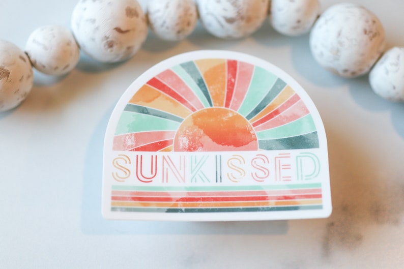 May include: A sticker featuring a watercolour-style illustration of a sun with a rainbow behind it. The text "SUNKISSED" is written in a retro font below the sun.