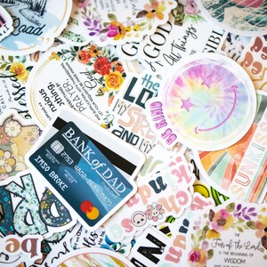 May include: A collection of colorful stickers with various designs and text, including floral patterns, inspirational quotes, and a tie-dye smiley face. A credit card with the text "BANK of DAD" is also visible.