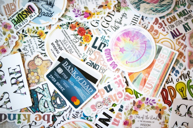 May include: A collection of colorful stickers with various designs and text, including floral patterns, inspirational quotes, and a tie-dye smiley face. A credit card with the text "BANK of DAD" is also visible.
