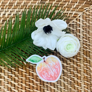 Op de afbeelding: Een sticker in perzikkleur met een aquarelontwerp van een perzik en de tekst "Just Peachy" in witte cursieve letters.
