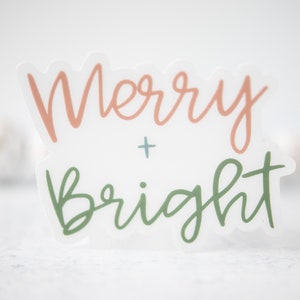 K&ouml;nnte beinhalten: Ein wei&szlig;er Aufkleber mit dem Text "Merry + Bright" in rosa und gr&uuml;ner Schrift.