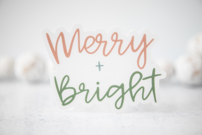 K&ouml;nnte beinhalten: Ein wei&szlig;er Aufkleber mit dem Text "Merry + Bright" in rosa und gr&uuml;ner Schrift.