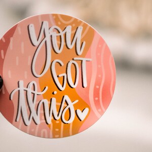 May include: A round sticker with an abstract design in shades of pink, orange, and white. The sticker features the text "You got this" in white script with a small pink heart.