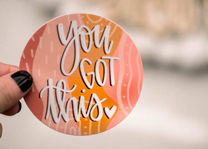 May include: A round sticker with an abstract design in shades of pink, orange, and white. The sticker features the text "You got this" in white script with a small pink heart.