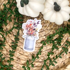 Pu&ograve; includere: Un adesivo con una lattina di latte vintage piena di un bouquet di fiori autunnali. I fiori sono un mix di arancione, rosso, giallo e viola.