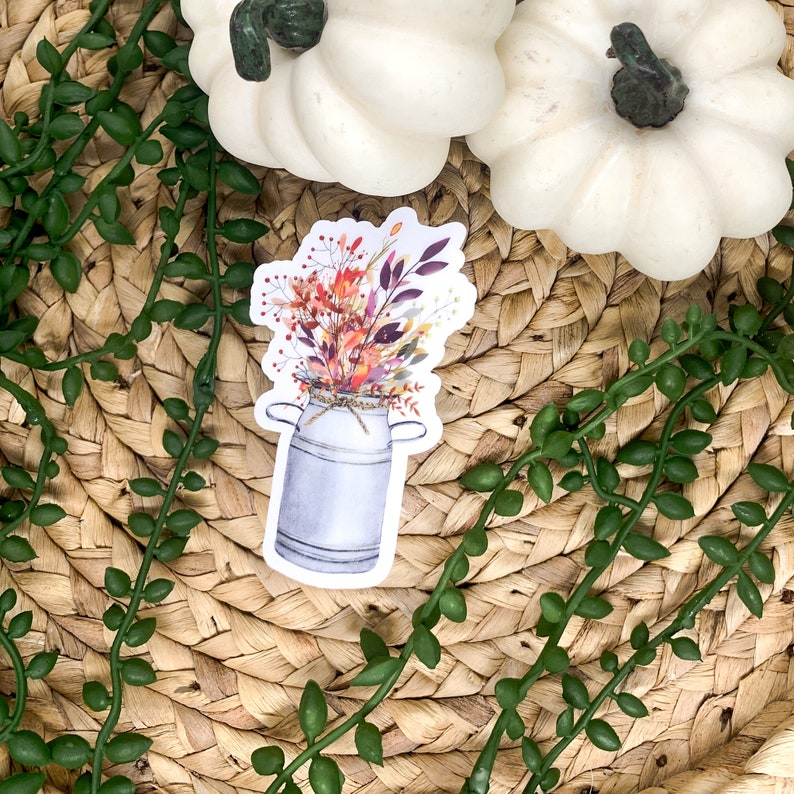 Pu&ograve; includere: Un adesivo con una lattina di latte vintage piena di un bouquet di fiori autunnali. I fiori sono un mix di arancione, rosso, giallo e viola.