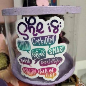 Puede incluir: Un tarro de vidrio con tapa morada y una pegatina que dice "Ella es: Fuerte, Confidente, Valiente, Inteligente, Feroz, Poderosa, Hermosa, Llena de Fuego".