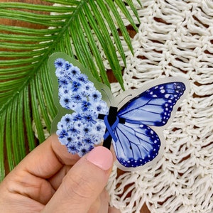 May include: A clear sticker with a blue butterfly design. The butterfly is made of blue flowers on one side and a blue butterfly with white spots on the other side. A blue ribbon is tied around the center of the butterfly.