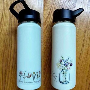 May include: Two white stainless steel water bottles with black lids and handles. One bottle has a sticker with a bouquet of flowers in a glass jar. The other bottle has a sticker with a row of flowers and the text "Grow Positive Thoughts".