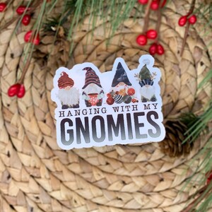 K&ouml;nnte beinhalten: Ein wei&szlig;er Aufkleber mit vier Gnomen, die rote, schwarze und wei&szlig;e M&uuml;tzen tragen. Die Gnome halten Weihnachtskugeln. Der Aufkleber lautet "Hanging with my Gnomies".