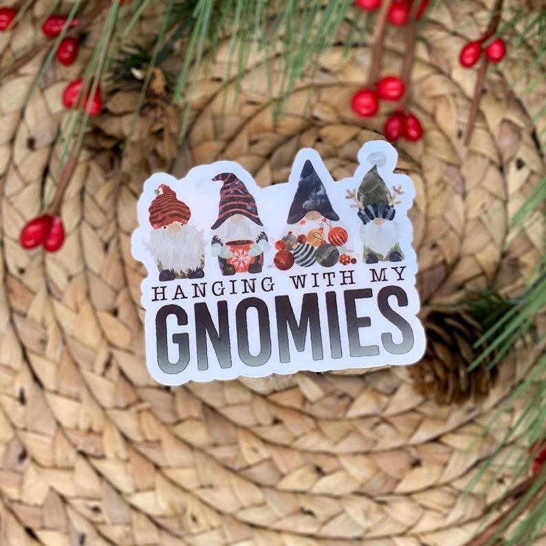 K&ouml;nnte beinhalten: Ein wei&szlig;er Aufkleber mit vier Gnomen, die rote, schwarze und wei&szlig;e M&uuml;tzen tragen. Die Gnome halten Weihnachtskugeln. Der Aufkleber lautet "Hanging with my Gnomies".