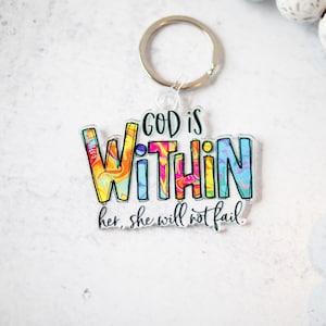 May include: A clear acrylic keychain with a silver keyring. The keychain features the words "GOD IS WITHIN" in colorful, bold lettering. Below the text is the phrase "her, she will not fail."