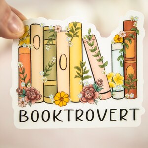 Pu&ograve; includere: Un adesivo bianco con una pila di libri colorati con accenti floreali e il testo "BOOKTROVERT" in nero.