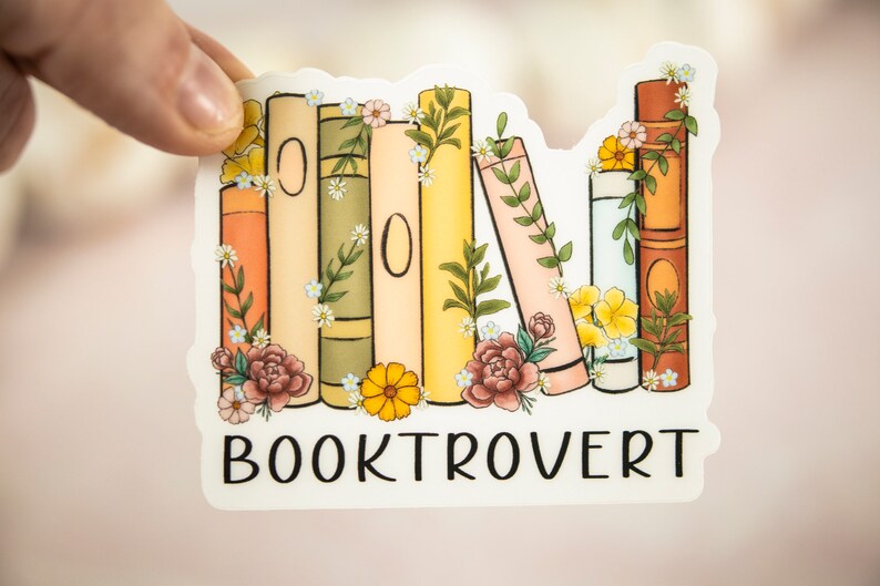 Pu&ograve; includere: Un adesivo bianco con una pila di libri colorati con accenti floreali e il testo "BOOKTROVERT" in nero.