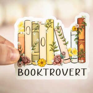 Pu&ograve; includere: Un adesivo con una pila di libri colorati con accenti floreali e il testo "BOOKTROVERT" sotto.