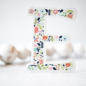May include: A white letter E with a floral pattern in shades of pink, blue, and green. The letter is standing on a white surface.