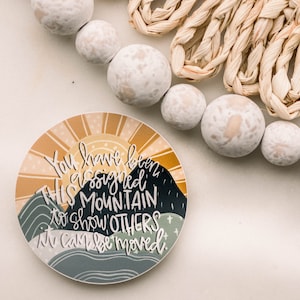 May include: A round sticker with a sunrise and mountains in the background. The text on the sticker reads "You have been assigned THIS MOUNTAIN to show OTHERS it can be moved."