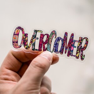 Puede incluir: Una pegatina transparente con la palabra "Overcomer" escrita en letras coloridas y estampadas.