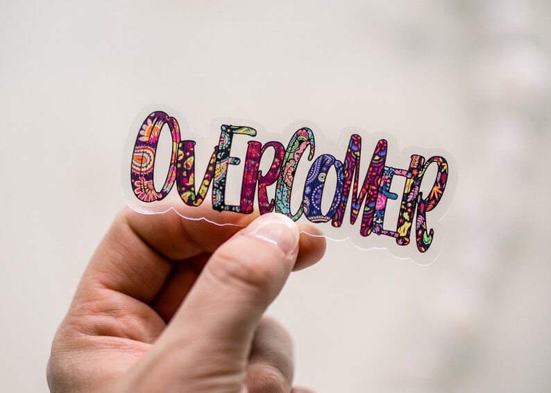 Puede incluir: Una pegatina transparente con la palabra "Overcomer" escrita en letras coloridas y estampadas.