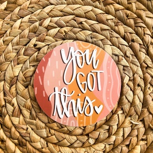 May include: A round sticker with an abstract pink and orange background. The sticker has white text that says "You got this" with a small pink heart.