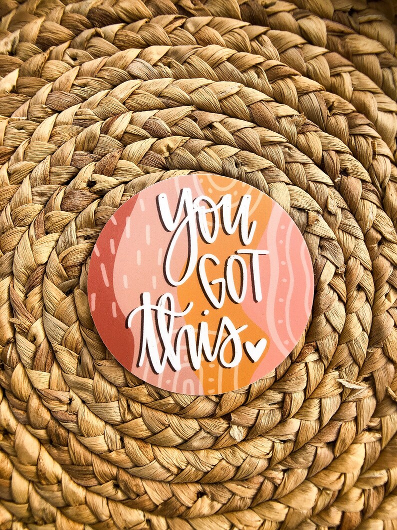 May include: A round sticker with an abstract pink and orange background. The sticker has white text that says "You got this" with a small pink heart.