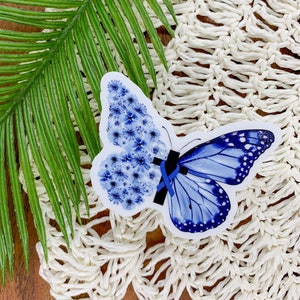 May include: A blue butterfly sticker with a blue ribbon. The butterfly is made of blue flowers. The sticker is on a white crocheted surface.