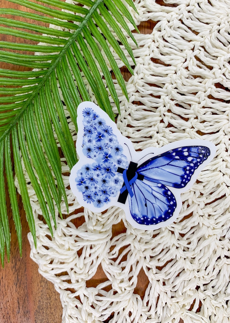 May include: A blue butterfly sticker with a blue ribbon. The butterfly is made of blue flowers. The sticker is on a white crocheted surface.