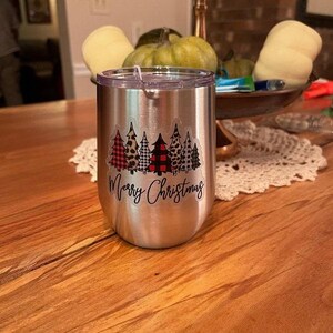 Puede incluir: Un vaso de acero inoxidable con una pegatina blanca que dice "Feliz Navidad" y presenta una fila de cinco &aacute;rboles de Navidad con diferentes patrones, incluyendo cuadros, estampado de leopardo y lunares.