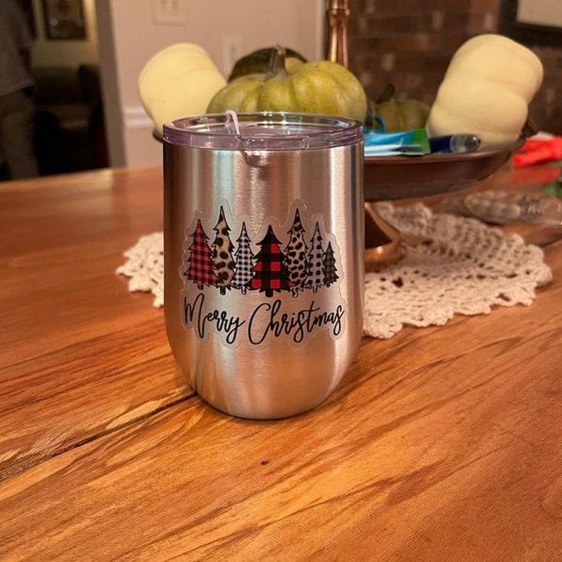 Puede incluir: Un vaso de acero inoxidable con una pegatina blanca que dice "Feliz Navidad" y presenta una fila de cinco &aacute;rboles de Navidad con diferentes patrones, incluyendo cuadros, estampado de leopardo y lunares.
