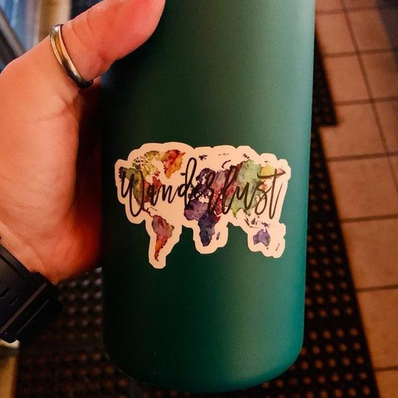 May include: A dark green water bottle with a white sticker that says "Wanderlust" in cursive over a colorful world map design. The bottle is held by a person wearing a ring.