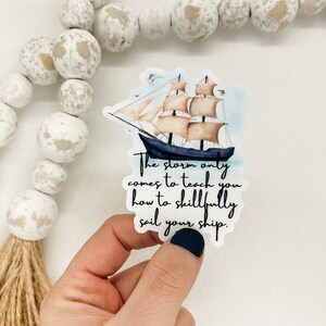 以下が含まれることがあります： 嵐の海を航海する船の水彩画が描かれたステッカー。テキストには「嵐は、あなたに巧みに船を操る方法を教えるためにやってくるだけだ。」と書いてあります。