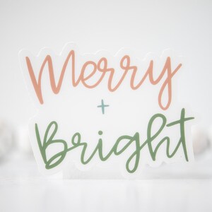 K&ouml;nnte beinhalten: Ein wei&szlig;er Aufkleber mit dem Text "Merry + Bright" in Kursivschrift. Das Wort "Merry" ist in Pfirsichfarbe und das Wort "Bright" in Gr&uuml;n.