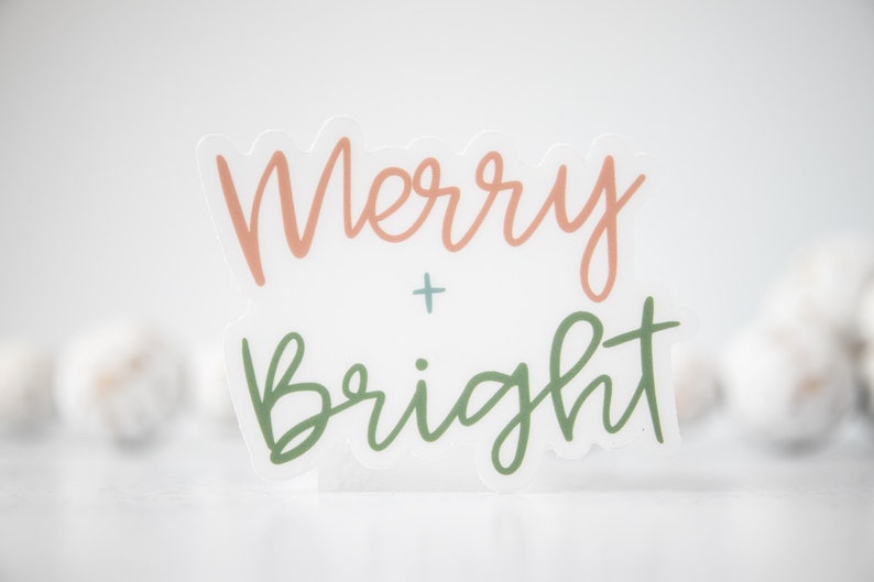 K&ouml;nnte beinhalten: Ein wei&szlig;er Aufkleber mit dem Text "Merry + Bright" in Kursivschrift. Das Wort "Merry" ist in Pfirsichfarbe und das Wort "Bright" in Gr&uuml;n.