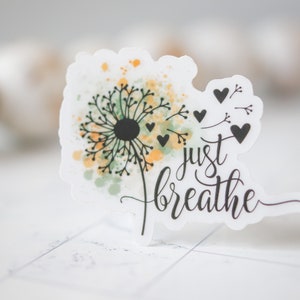 Puede incluir: Una pegatina blanca con un dise&ntilde;o de diente de le&oacute;n acuarela y el texto "just breathe". El diente de le&oacute;n tiene acentos acuarela verde y amarillo y semillas negras. Hay peque&ntilde;os corazones negros alrededor del diente de le&oacute;n.