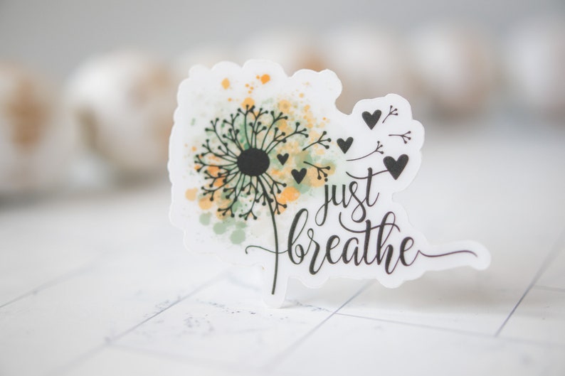 Puede incluir: Una pegatina blanca con un dise&ntilde;o de diente de le&oacute;n acuarela y el texto "just breathe". El diente de le&oacute;n tiene acentos acuarela verde y amarillo y semillas negras. Hay peque&ntilde;os corazones negros alrededor del diente de le&oacute;n.