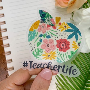 May include: A clear sticker in the shape of an apple, filled with a floral pattern in pink, yellow, and teal. The sticker has the text "#TeacherLife" in dark gray.