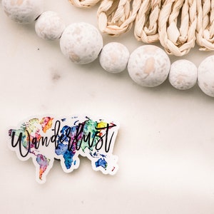 May include: A white sticker with a watercolor world map design and the word "Wanderlust" in black script. The sticker is on a white surface, with white beaded garland and woven raffia accents in the background.