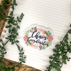 May include: A white sticker with a floral wreath design. The wreath is made up of pink, orange, and green flowers. The text "Chaos Coordinator" is written in black cursive lettering inside the wreath.