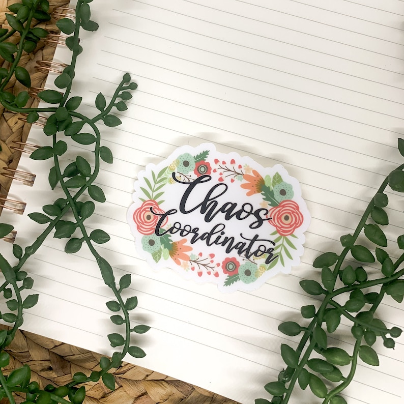 May include: A white sticker with a floral wreath design. The wreath is made up of pink, orange, and green flowers. The text "Chaos Coordinator" is written in black cursive lettering inside the wreath.