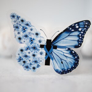 May include: A blue butterfly with a cross and a blue ribbon on its body. The butterfly is made of clear plastic and has a floral design on one wing.
